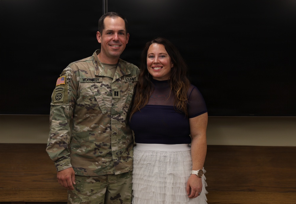 A U.S. Army soldier in OCP (Operational Camouflage Pattern) uniform stands alongside a civilian woman, both smiling for a posed photograph. The soldier's name tape reads 'MCKENNEDY' and displays a Captain rank insignia (two vertical bars) on the chest, along with an 82nd Airborne Division patch and Airborne tab on the left sleeve. The setting appears to be an indoor facility with a dark chalkboard or presentation board in the background.
