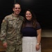 A U.S. Army soldier in OCP (Operational Camouflage Pattern) uniform stands alongside a civilian woman, both smiling for a posed photograph. The soldier's name tape reads 'MCKENNEDY' and displays a Captain rank insignia (two vertical bars) on the chest, along with an 82nd Airborne Division patch and Airborne tab on the left sleeve. The setting appears to be an indoor facility with a dark chalkboard or presentation board in the background.