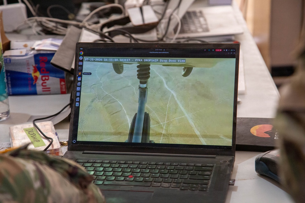 A Lenovo ThinkPad laptop displays a live or recorded camera feed labeled 'PYKA DROPSHIP Drop Door View' dated 07-21-2026, showing an aerial downward-looking camera view of agricultural fields. The workstation appears to be an operations or ground control station, cluttered with cables, a Red Bull can, and other items. The screen suggests drone or autonomous aircraft monitoring operations.