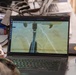 A Lenovo ThinkPad laptop displays a live or recorded camera feed labeled 'PYKA DROPSHIP Drop Door View' dated 07-21-2026, showing an aerial downward-looking camera view of agricultural fields. The workstation appears to be an operations or ground control station, cluttered with cables, a Red Bull can, and other items. The screen suggests drone or autonomous aircraft monitoring operations.
