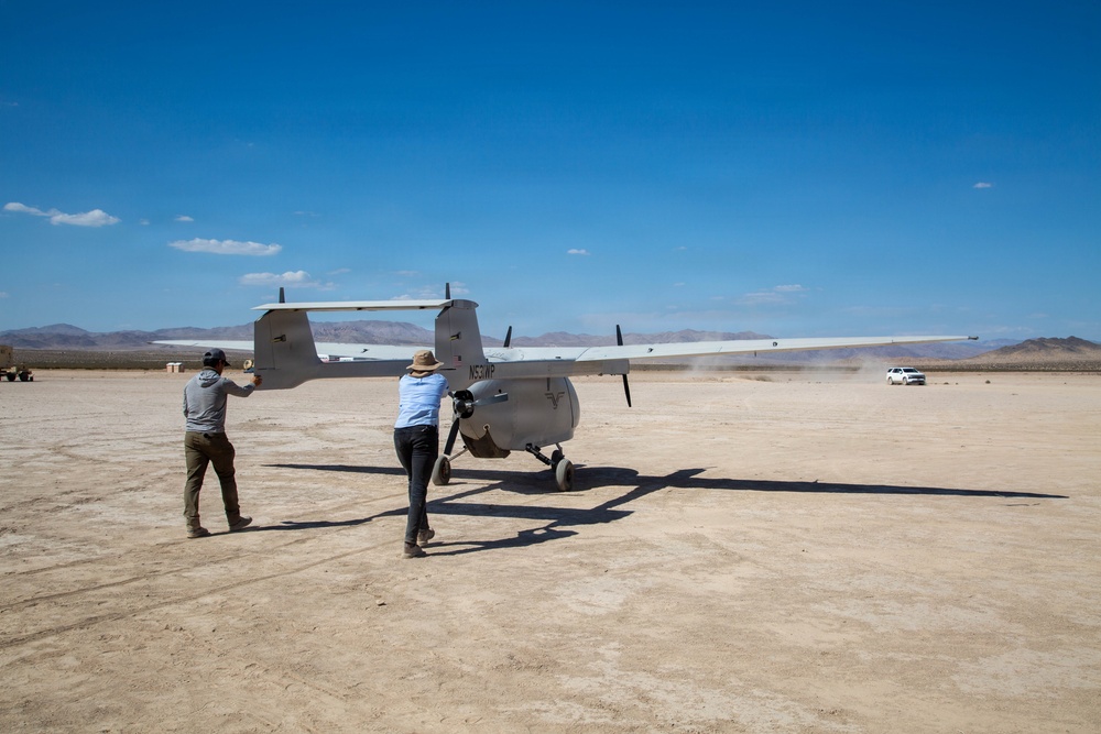 Two individuals are working with a small propeller-driven aircraft on a dry desert lakebed or airstrip under clear blue skies. The aircraft, bearing the registration number N53WP, appears to be a twin-boom pusher-configuration unmanned or light manned aircraft. A white SUV is visible in the background, and mountains line the distant horizon.