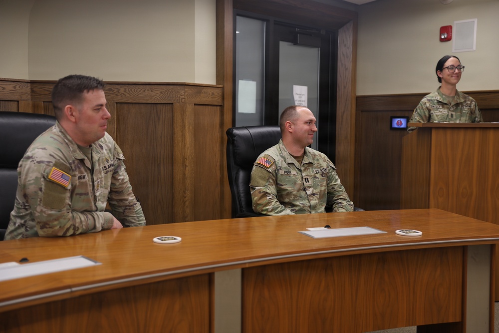 Three U.S. Army soldiers are gathered in what appears to be a formal military briefing or ceremony room. Two male soldiers are seated at a wooden table while a female soldier stands at a podium, apparently speaking or presenting. The setting resembles a courtroom or formal conference room with wood paneling.