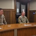 Three U.S. Army soldiers are gathered in what appears to be a formal military briefing or ceremony room. Two male soldiers are seated at a wooden table while a female soldier stands at a podium, apparently speaking or presenting. The setting resembles a courtroom or formal conference room with wood paneling.