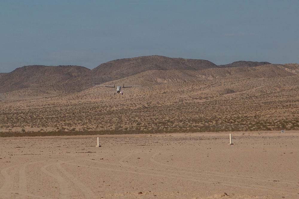 A small aircraft is captured at low altitude over a desert landscape, appearing to be either landing or taking off from an unpaved desert airstrip. The terrain is arid and flat in the foreground, marked by white runway marker posts, with sparse desert scrub vegetation and rocky hills in the background. The sky is clear with a slight haze.
