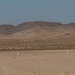 A small aircraft is captured at low altitude over a desert landscape, appearing to be either landing or taking off from an unpaved desert airstrip. The terrain is arid and flat in the foreground, marked by white runway marker posts, with sparse desert scrub vegetation and rocky hills in the background. The sky is clear with a slight haze.