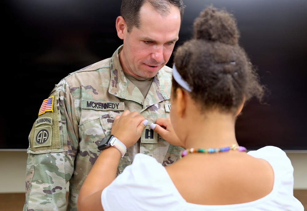 A U.S. Army soldier named McKennedy, wearing an OCP (Operational Camouflage Pattern) uniform with 82nd Airborne Division and Airborne patches, is having an insignia or rank device pinned onto his chest by a young girl. The scene appears to be a military promotion or award ceremony. The soldier is looking down attentively as the girl performs the pinning.