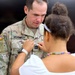 A U.S. Army soldier named McKennedy, wearing an OCP (Operational Camouflage Pattern) uniform with 82nd Airborne Division and Airborne patches, is having an insignia or rank device pinned onto his chest by a young girl. The scene appears to be a military promotion or award ceremony. The soldier is looking down attentively as the girl performs the pinning.