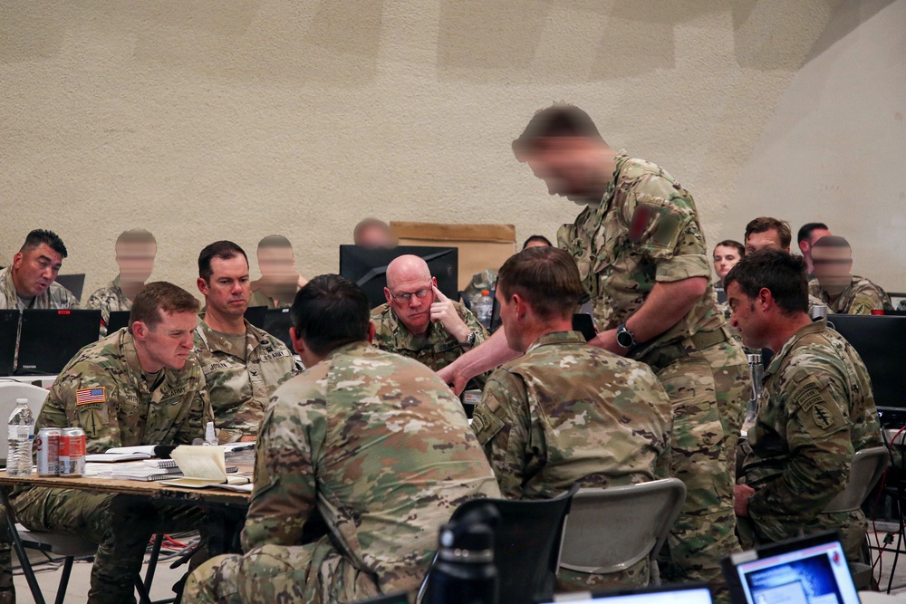 A group of U.S. Army soldiers in Operational Camouflage Pattern (OCP) uniforms are gathered around a table in what appears to be a military operations or planning room. Several personnel are engaged in an apparent briefing or discussion, with laptops, notebooks, and documents visible on the table. The setting suggests a command post, training exercise, or operational planning session.