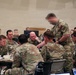 A group of U.S. Army soldiers in Operational Camouflage Pattern (OCP) uniforms are gathered around a table in what appears to be a military operations or planning room. Several personnel are engaged in an apparent briefing or discussion, with laptops, notebooks, and documents visible on the table. The setting suggests a command post, training exercise, or operational planning session.