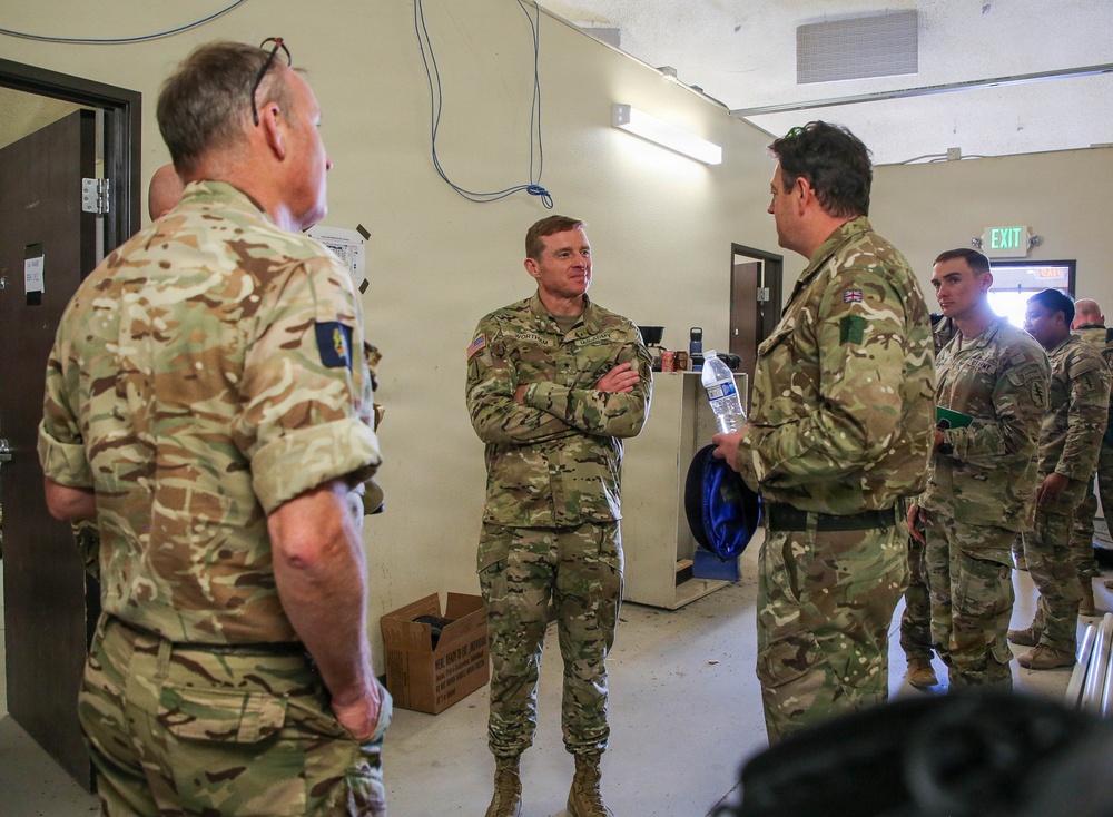 A group of military personnel from at least two different nations are gathered indoors in what appears to be a military facility or briefing room. The central figure, a U.S. Army soldier with arms crossed, appears to be engaged in conversation with British Army soldiers wearing MTP (Multi-Terrain Pattern) camouflage uniforms. The setting suggests an inter-allied meeting, briefing, or coordination session.