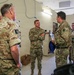 A group of military personnel from at least two different nations are gathered indoors in what appears to be a military facility or briefing room. The central figure, a U.S. Army soldier with arms crossed, appears to be engaged in conversation with British Army soldiers wearing MTP (Multi-Terrain Pattern) camouflage uniforms. The setting suggests an inter-allied meeting, briefing, or coordination session.