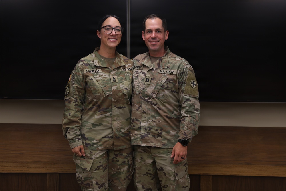 Two U.S. Army soldiers in Operational Camouflage Pattern (OCP) uniforms are posing together for a photo indoors. The soldier on the left, identified by name tape as 'CONNELLY,' appears to hold a senior NCO rank, while the soldier on the right, identified as 'MOSEN,' displays a Captain's rank insignia and a medical cross patch. Both are smiling and standing in front of a blackboard.