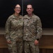Two U.S. Army soldiers in Operational Camouflage Pattern (OCP) uniforms are posing together for a photo indoors. The soldier on the left, identified by name tape as 'CONNELLY,' appears to hold a senior NCO rank, while the soldier on the right, identified as 'MOSEN,' displays a Captain's rank insignia and a medical cross patch. Both are smiling and standing in front of a blackboard.