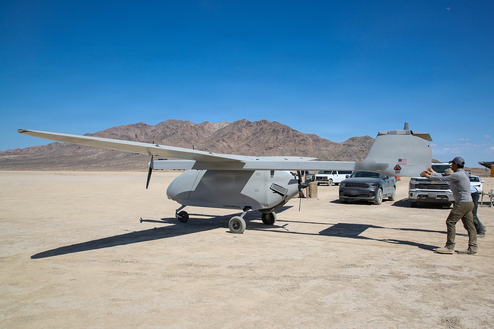 A gray unmanned aerial vehicle (UAV) or experimental aircraft is parked on a desert dry lakebed, with the word 'EXPERIMENTAL' visible on its fuselage. A person appears to be photographing or inspecting the aircraft, while several pickup trucks and support vehicles are parked nearby. The setting appears to be a remote desert test site, likely in the American Southwest based on the terrain and mountain backdrop.