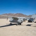 A gray unmanned aerial vehicle (UAV) or experimental aircraft is parked on a desert dry lakebed, with the word 'EXPERIMENTAL' visible on its fuselage. A person appears to be photographing or inspecting the aircraft, while several pickup trucks and support vehicles are parked nearby. The setting appears to be a remote desert test site, likely in the American Southwest based on the terrain and mountain backdrop.