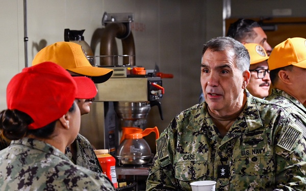 A U.S. Navy officer with the name tape 'CASE' and a single star insignia (indicating the rank of Rear Admiral Lower Half/Brigadier General equivalent) is conversing with enlisted personnel in what appears to be a galley or mess facility aboard a naval vessel or installation. The officer is holding a white disposable cup while engaging with sailors wearing red and orange baseball caps, which are typical of color-coded work center or division designations. The setting suggests an informal visit or inspection of food service operations.
