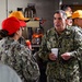 A U.S. Navy officer with the name tape 'CASE' and a single star insignia (indicating the rank of Rear Admiral Lower Half/Brigadier General equivalent) is conversing with enlisted personnel in what appears to be a galley or mess facility aboard a naval vessel or installation. The officer is holding a white disposable cup while engaging with sailors wearing red and orange baseball caps, which are typical of color-coded work center or division designations. The setting suggests an informal visit or inspection of food service operations.