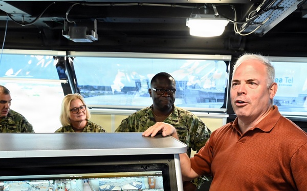 A civilian man in a brown polo shirt appears to be briefing or presenting to a group of U.S. Navy personnel inside what appears to be a ship's bridge or command center. A large multi-panel surveillance monitor displaying numerous camera feeds is prominently visible in the foreground. The setting appears to be aboard a naval vessel, with windows showing a waterfront or port environment in the background.