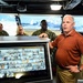 A civilian man in a brown polo shirt appears to be briefing or presenting to a group of U.S. Navy personnel inside what appears to be a ship's bridge or command center. A large multi-panel surveillance monitor displaying numerous camera feeds is prominently visible in the foreground. The setting appears to be aboard a naval vessel, with windows showing a waterfront or port environment in the background.
