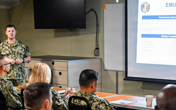 A U.S. Navy service member is delivering a briefing on 'EMU Core Capabilities' to a group of fellow Navy personnel seated at tables in a conference room. The presentation slide visible on the projected screen includes sections on 'Performance Specifications' and 'Medical Capabilities.' All individuals are wearing Navy Working Uniform (NWU) Type III camouflage.