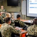 A U.S. Navy service member is delivering a briefing on 'EMU Core Capabilities' to a group of fellow Navy personnel seated at tables in a conference room. The presentation slide visible on the projected screen includes sections on 'Performance Specifications' and 'Medical Capabilities.' All individuals are wearing Navy Working Uniform (NWU) Type III camouflage.