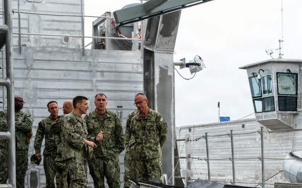 A military training or demonstration exercise is taking place on what appears to be a naval vessel or dock facility. A person in desert camouflage uniform is strapped to a litter/stretcher in the foreground, simulating a casualty. Several uniformed Navy personnel in NWU (Navy Working Uniform) camouflage are observing and discussing in the background.