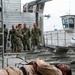 A military training or demonstration exercise is taking place on what appears to be a naval vessel or dock facility. A person in desert camouflage uniform is strapped to a litter/stretcher in the foreground, simulating a casualty. Several uniformed Navy personnel in NWU (Navy Working Uniform) camouflage are observing and discussing in the background.