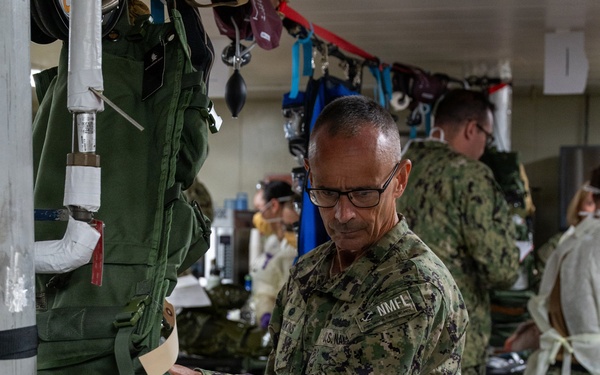 A U.S. Navy personnel member in NWU Type III camouflage uniform is working in what appears to be a military medical training or triage facility. The scene shows multiple personnel engaged in medical simulation activities, with a simulated casualty visible in the foreground displaying mock wounds and wearing gold athletic shoes. The environment contains extensive medical equipment, supplies, and training props consistent with a mass casualty exercise.