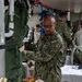 A U.S. Navy personnel member in NWU Type III camouflage uniform is working in what appears to be a military medical training or triage facility. The scene shows multiple personnel engaged in medical simulation activities, with a simulated casualty visible in the foreground displaying mock wounds and wearing gold athletic shoes. The environment contains extensive medical equipment, supplies, and training props consistent with a mass casualty exercise.