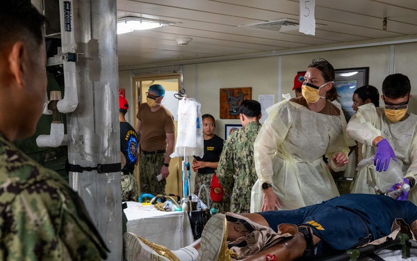 A military medical training or casualty drill scenario taking place in what appears to be a shipboard medical bay or military medical facility. A simulated casualty in military uniform is lying on a stretcher/litter with mock wound moulage visible on the leg, while medical personnel in PPE (gowns, masks, gloves) attend to the patient. Additional military personnel in camouflage uniforms observe the exercise.