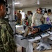 A military medical training or casualty drill scenario taking place in what appears to be a shipboard medical bay or military medical facility. A simulated casualty in military uniform is lying on a stretcher/litter with mock wound moulage visible on the leg, while medical personnel in PPE (gowns, masks, gloves) attend to the patient. Additional military personnel in camouflage uniforms observe the exercise.