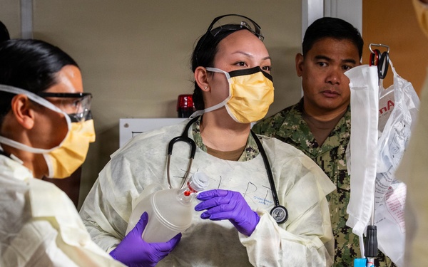 A medical team in full personal protective equipment is attending to a patient lying on a gurney or bed. At least two personnel are wearing yellow isolation gowns, purple nitrile gloves, face masks, and safety glasses, with one wearing a stethoscope and holding a medical cup or container. A person in military camouflage uniform observes in the background, suggesting a military medical setting.