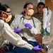 A medical team in full personal protective equipment is attending to a patient lying on a gurney or bed. At least two personnel are wearing yellow isolation gowns, purple nitrile gloves, face masks, and safety glasses, with one wearing a stethoscope and holding a medical cup or container. A person in military camouflage uniform observes in the background, suggesting a military medical setting.