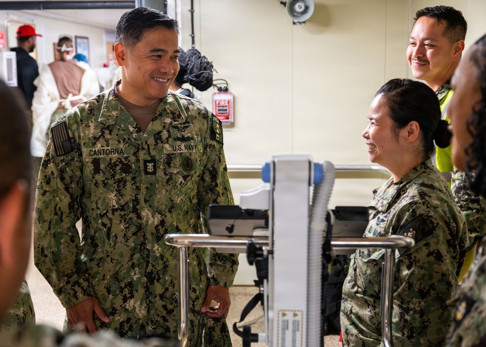 A U.S. Navy servicemember named Cantorna, wearing NWU Type III digital camouflage, is engaged in a friendly conversation with fellow Navy personnel in what appears to be a medical or training facility. Multiple uniformed personnel are present, along with civilians visible in the background. The atmosphere appears relaxed and collegial.