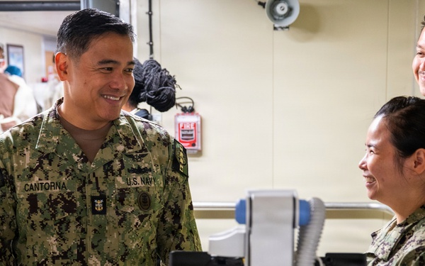 A U.S. Navy servicemember named Cantorna, wearing NWU Type III digital camouflage, is engaged in a friendly conversation with fellow Navy personnel in what appears to be a medical or training facility. Multiple uniformed personnel are present, along with civilians visible in the background. The atmosphere appears relaxed and collegial.