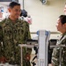 A U.S. Navy servicemember named Cantorna, wearing NWU Type III digital camouflage, is engaged in a friendly conversation with fellow Navy personnel in what appears to be a medical or training facility. Multiple uniformed personnel are present, along with civilians visible in the background. The atmosphere appears relaxed and collegial.