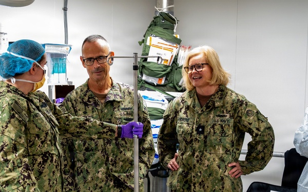 The image shows a group of U.S. Navy personnel in digital camouflage uniforms gathered in what appears to be a military medical facility or field hospital. Three individuals in Navy working uniforms are engaged in conversation near a medical gurney or examination table, while two others in medical protective gear (gowns, gloves, and hair covers) are also present. The setting contains visible medical equipment and supplies consistent with a clinical or surgical environment.