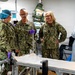 The image shows a group of U.S. Navy personnel in digital camouflage uniforms gathered in what appears to be a military medical facility or field hospital. Three individuals in Navy working uniforms are engaged in conversation near a medical gurney or examination table, while two others in medical protective gear (gowns, gloves, and hair covers) are also present. The setting contains visible medical equipment and supplies consistent with a clinical or surgical environment.