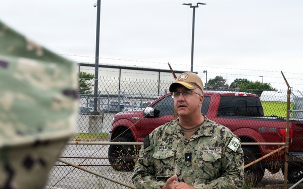 A U.S. Navy officer in digital camouflage (NWU Type III) uniform is addressing a small group of people outdoors near a secured fenced area. The officer's name tape reads 'BRAJEVIC' and displays a single star insignia indicating the rank of Rear Admiral (Lower Half) or Brigadier General equivalent. The setting appears to be a military installation or industrial facility.