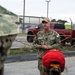 A U.S. Navy officer in digital camouflage (NWU Type III) uniform is addressing a small group of people outdoors near a secured fenced area. The officer's name tape reads 'BRAJEVIC' and displays a single star insignia indicating the rank of Rear Admiral (Lower Half) or Brigadier General equivalent. The setting appears to be a military installation or industrial facility.
