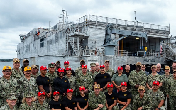A large group of approximately 40 military personnel pose for a group photo on a pier in front of a U.S. Naval vessel. The ship visible in the background is identified as 'US Naval Ship Point Loma' with the name 'Appleton' also visible on an upper structure. Personnel are wearing a mix of Navy Working Uniform (NWU) camouflage, black uniforms, and colored caps (red, gold, and tan).