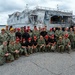 A large group of approximately 40 military personnel pose for a group photo on a pier in front of a U.S. Naval vessel. The ship visible in the background is identified as 'US Naval Ship Point Loma' with the name 'Appleton' also visible on an upper structure. Personnel are wearing a mix of Navy Working Uniform (NWU) camouflage, black uniforms, and colored caps (red, gold, and tan).