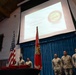 A U.S. Marine Corps Corporal's Course Graduation ceremony is taking place in a formal hall or chapel setting. Several Marines in camouflage utility uniforms stand at attention on a stage in front of a blue curtain backdrop. A projected screen displays 'Welcome to Corporal's Course Graduation' along with a Task Force Forge unit patch, and red diploma folders are stacked on a table.