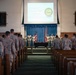 A U.S. Marine Corps Corporal's Course Graduation ceremony is taking place in what appears to be a chapel or auditorium-style venue. Marines in desert camouflage uniforms (MARPAT) are standing at attention in rows facing a stage, where a group of graduates and officials stand before an American flag and Marine Corps flag. A projection screen displays 'Welcome to Corporal's Course Graduation' along with a military unit seal.