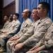 A row of military personnel in matching camouflage uniforms are seated in chairs against a blue curtain backdrop, appearing to be at a formal ceremony or official event. The group consists of approximately seven individuals of diverse backgrounds, including both male and female service members. Their attire and posture suggest a structured, formal military setting.