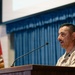 A U.S. Marine in desert MARPAT camouflage uniform is speaking at a wooden podium equipped with a microphone. The setting appears to be a formal military briefing or ceremony held in an auditorium or conference room. A red and gold military flag or guidon is visible in the background alongside blue curtains and a projection screen.