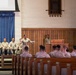 A military ceremony or briefing is taking place inside what appears to be a chapel or auditorium on a military installation. A service member in desert camouflage uniform is addressing an audience of seated military personnel from the front of the room, while additional personnel are seated on a stage area alongside a military flag. The setting, pew-style seating, and formal arrangement suggest an official military ceremony such as an awards presentation or change of command.