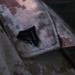 The image shows a close-up of a military uniform collar or epaulette area featuring a black metal rank insignia. The uniform displays a digital camouflage pattern consistent with USMC MARPAT (Marine Pattern) desert camouflage. The rank insignia appears to be a Corporal (E-4) chevron in subdued black finish.