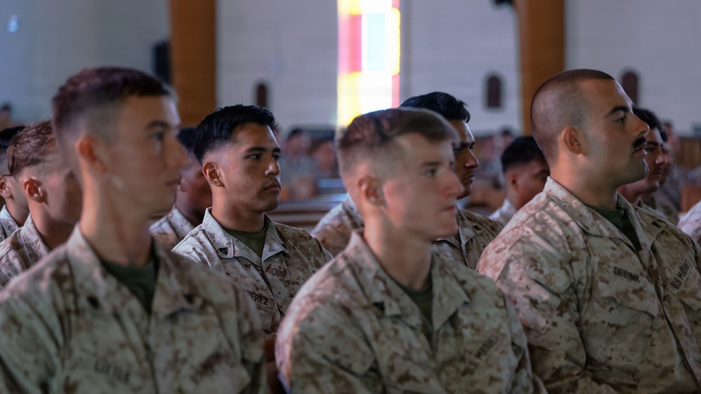 A group of U.S. Marines in MARPAT camouflage uniforms are seated in rows inside what appears to be a chapel or large assembly hall. They appear to be attending a formal ceremony or briefing, facing attentively in the same direction. Multiple rows of personnel are visible extending into the background.