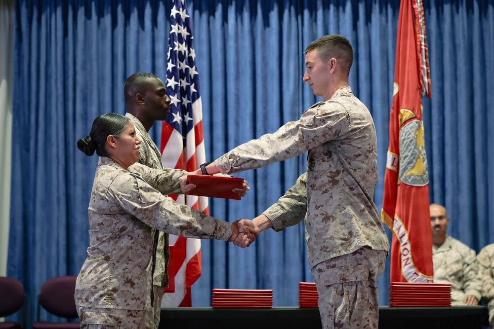A military ceremony is taking place in an indoor venue where a Marine in desert MARPAT camouflage uniform is receiving a red folder/certificate from a female Marine while shaking hands. A second male Marine stands behind the presenting officer, and a third Marine is seated in the background. The U.S. flag and a Marine Corps unit flag are displayed on either side of the backdrop.